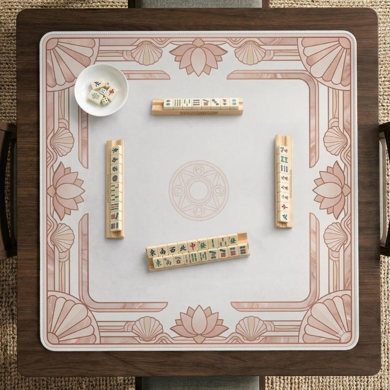 May include: A square mahjong table with a white playing surface and a decorative pink border featuring lotus flowers and seashells. The table includes mahjong tiles and a small bowl with dice.