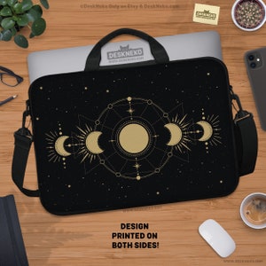 Puede incluir: Funda negra para portátil con un diseño celestial dorado de lunas, estrellas y soles. El texto "¡DISEÑO IMPRESO EN AMBOS LADOS!" es visible. La funda tiene un asa y una correa para el hombro. Sobre una superficie de madera.