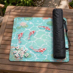 May include: A rectangular mahjong mat featuring a light blue water surface with white ripples, four red and white koi fish, green lily pads, and a pink lotus flower. Seven white mahjong tiles are placed on the mat, alongside a black carrying bag.
