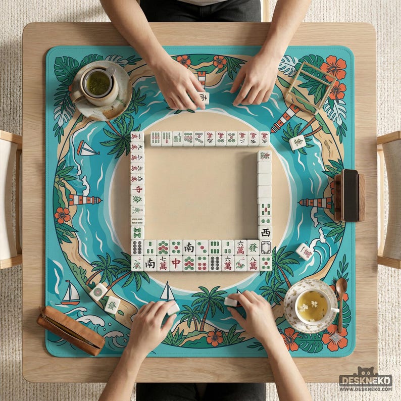 May include: A wooden table with a Mahjong game in play, featuring a tropical design. The board has a turquoise ocean scene with palm trees, sailboats, and a lighthouse. Players are arranging tiles, with cups of tea on the table.