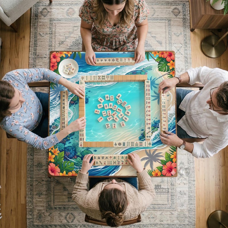 May include: A square mahjong table featuring a tropical beach print with blue water, sand, and hibiscus flowers. The table surface displays a central pile of mahjong tiles, with four racks of tiles arranged in a square formation for gameplay.