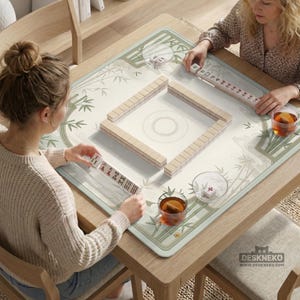 May include: A square mahjong mat featuring a light green and white bamboo pattern, placed on a wooden table. The mat displays a central square arrangement of ivory-colored mahjong tiles, with additional tiles held by players and a logo reading DESKNEKO.