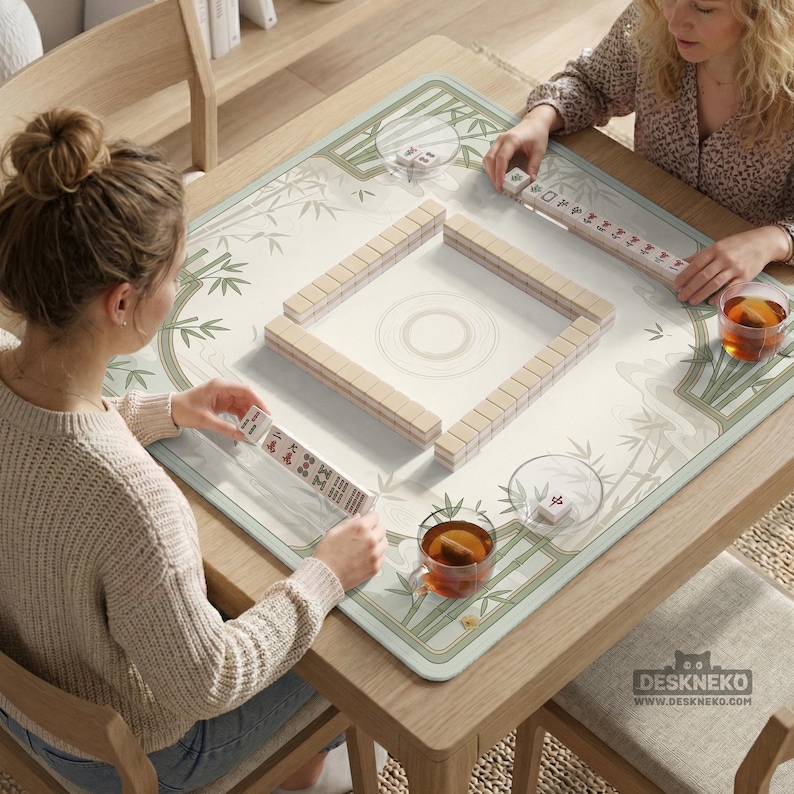 May include: A square mahjong mat featuring a light green and white bamboo pattern, placed on a wooden table. The mat displays a central square arrangement of ivory-colored mahjong tiles, with additional tiles held by players and a logo reading DESKNEKO.
