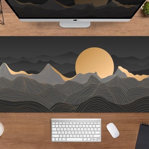 May include: A large, rectangular desk mat featuring a mountain landscape design. The design includes a golden sun, gray mountains, and gold accent lines on a dark gray background. A keyboard, mouse, and coffee cup are visible on the wooden desk.