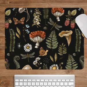 Puede incluir: Un ratón negro con un diseño de acuarela de setas, helechos, libélulas, margaritas y un caracol. Un ratón y un teclado de ordenador blancos son visibles sobre un escritorio de madera. La alfombrilla del ratón tiene forma rectangular.