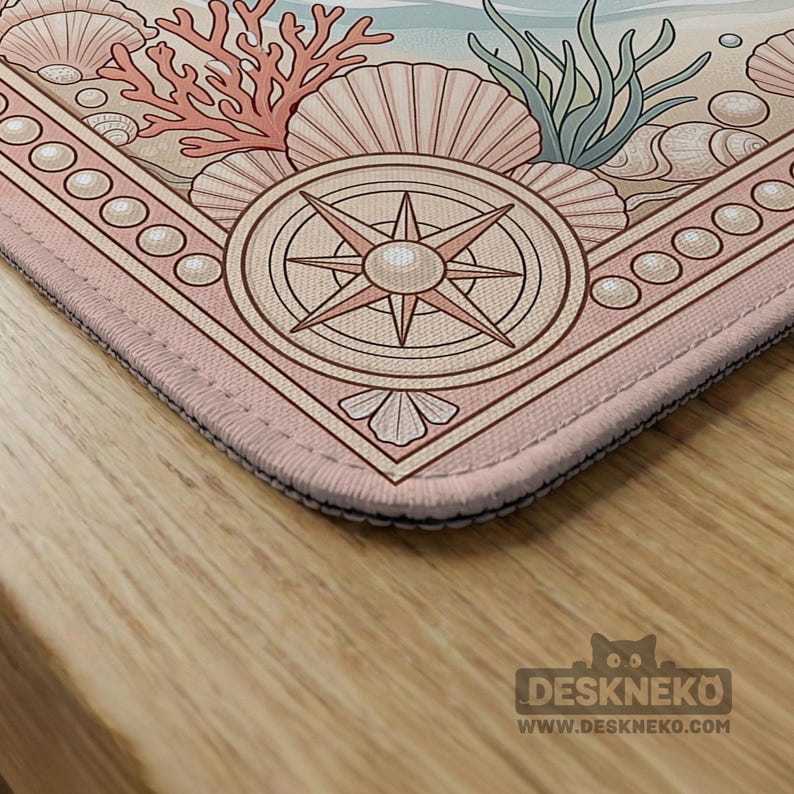 May include: A rectangular desk mat with a nautical design. The mat features a compass rose, seashells, coral, and seaweed in shades of pink, beige, and blue. The mat has a pink border and the text "DESKNEKO" and "WWW.DESKNEKO.COM".