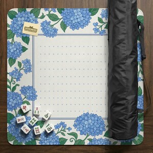May include: A white and blue floral play mat with a hydrangea design. The mat has a dotted center and a blue border. A black carrying case is rolled up on the right side. Several Mahjong tiles are scattered on the mat. The mat has the text "DESKNEKO".