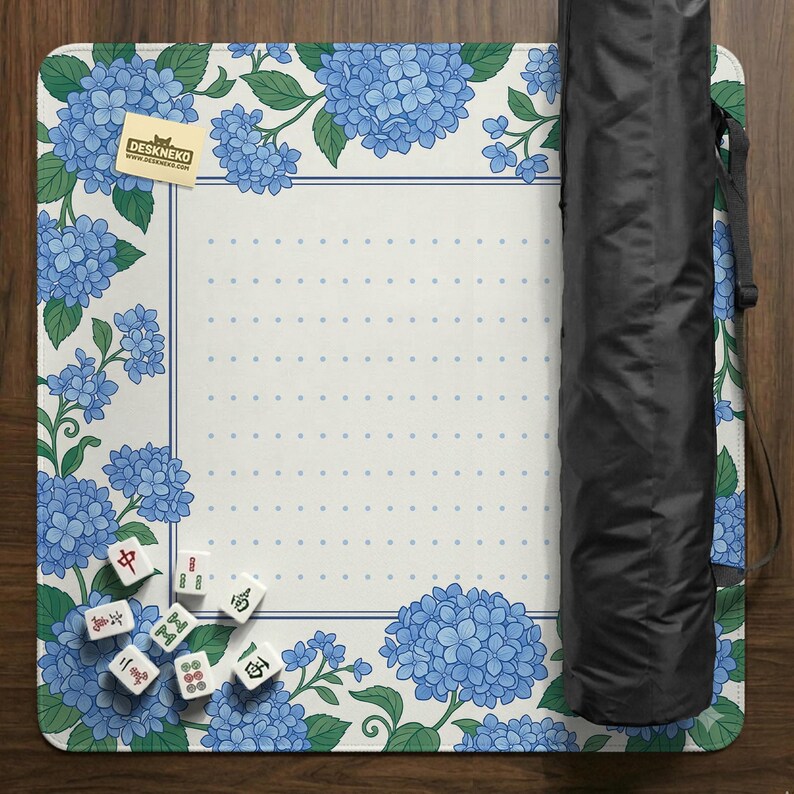 May include: A white and blue floral play mat with a hydrangea design. The mat has a dotted center and a blue border. A black carrying case is rolled up on the right side. Several Mahjong tiles are scattered on the mat. The mat has the text "DESKNEKO".