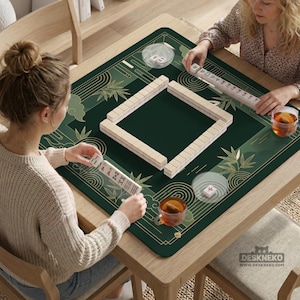 Pode incluir: Um tapete de jogo de Mahjong verde e dourado com desenhos de bambu e nuvens. O tapete est&aacute; sobre uma mesa de madeira com duas pessoas jogando Mahjong. O jogo inclui pe&ccedil;as e x&iacute;caras de ch&aacute;. O endere&ccedil;o do site "DESKNEKO" &eacute; vis&iacute;vel.