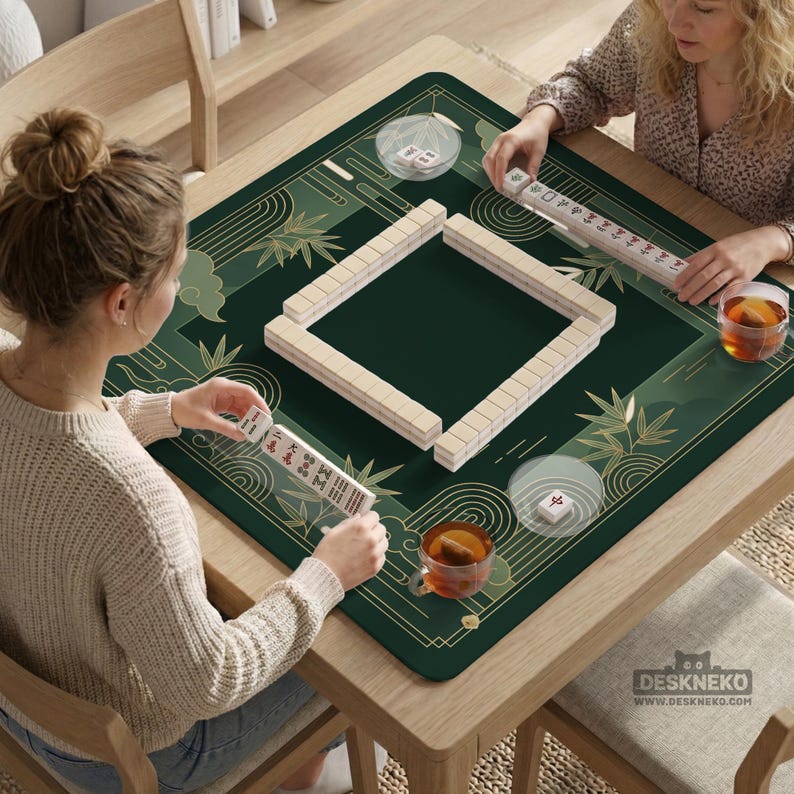 Pode incluir: Um tapete de jogo de Mahjong verde e dourado com desenhos de bambu e nuvens. O tapete est&aacute; sobre uma mesa de madeira com duas pessoas jogando Mahjong. O jogo inclui pe&ccedil;as e x&iacute;caras de ch&aacute;. O endere&ccedil;o do site "DESKNEKO" &eacute; vis&iacute;vel.
