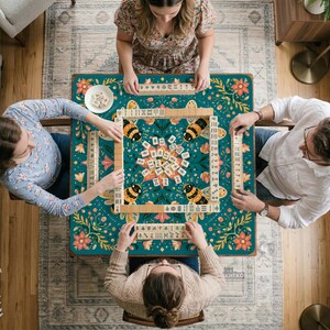May include: A square mahjong table cover featuring a dark teal background with a vibrant floral pattern and four large bumblebees in the corners. The center displays a collection of mahjong tiles, with additional tiles arranged in racks.