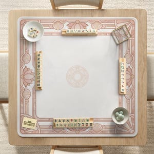 May include: A square wooden mahjong table with a white and pink decorative mat. The mat features a floral and geometric design. Small bowls with mahjong tiles and green balls are placed on the table. The logo "DESKNEKO" is visible.