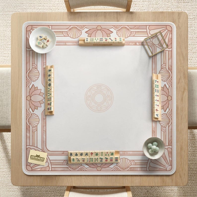 May include: A square wooden mahjong table with a white and pink decorative mat. The mat features a floral and geometric design. Small bowls with mahjong tiles and green balls are placed on the table. The logo "DESKNEKO" is visible.