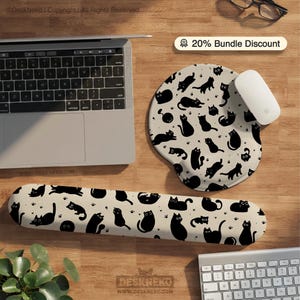 May include: A beige mouse mat and wrist rest set featuring a black cat design. The wrist rest is long and rectangular. The mouse mat is oval. A laptop, mouse, and keyboard are also visible on a wooden surface.