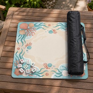 May include: A square mahjong mat featuring a light beige center surrounded by a border of teal seaweed, coral, seashells, and white pearls. A black cylindrical carrying case with a shoulder strap rests on the right side of the mat.