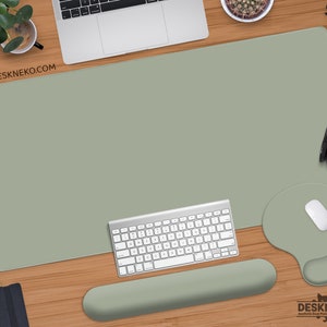 Puede incluir: Alfombrilla de escritorio verde salvia con un ratón y reposamuñecas a juego. Un teclado y un ratón blancos están sobre la alfombrilla. La dirección del sitio web www.deskneko.com es visible. La alfombrilla de escritorio está sobre una superficie de madera.