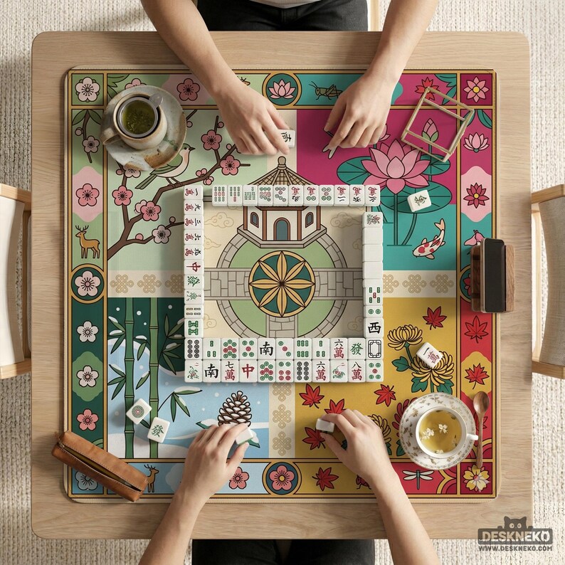 May include: A colorful Mahjong game in progress on a wooden table. The game board features a floral and nature-themed design with a pagoda, koi fish, and bamboo. Players are arranging tiles, with cups of tea nearby.