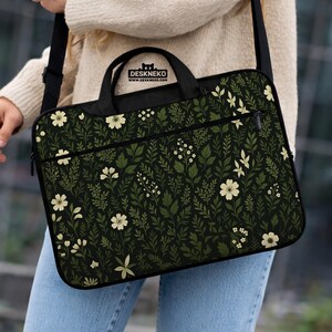 May include: Black laptop bag with a floral pattern of white flowers and green leaves. The bag has a black handle and shoulder strap. The text "DESKNEKO" is printed on the top of the bag.