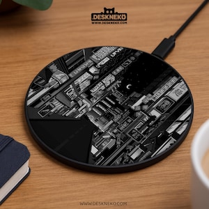 Puede incluir: Cargador inalámbrico negro con una detallada ilustración en escala de grises de un paisaje urbano nocturno, con edificios, letreros y una luna creciente. El cargador tiene un diseño elegante y redondo y un cable de carga negro.