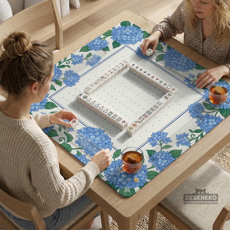 May include: A square wooden table with a floral Mahjong mat featuring blue hydrangea blooms. The mat surrounds a set of Mahjong tiles. Two people are playing Mahjong, with cups of tea on the table. The table has a "DESKNEKO" logo.