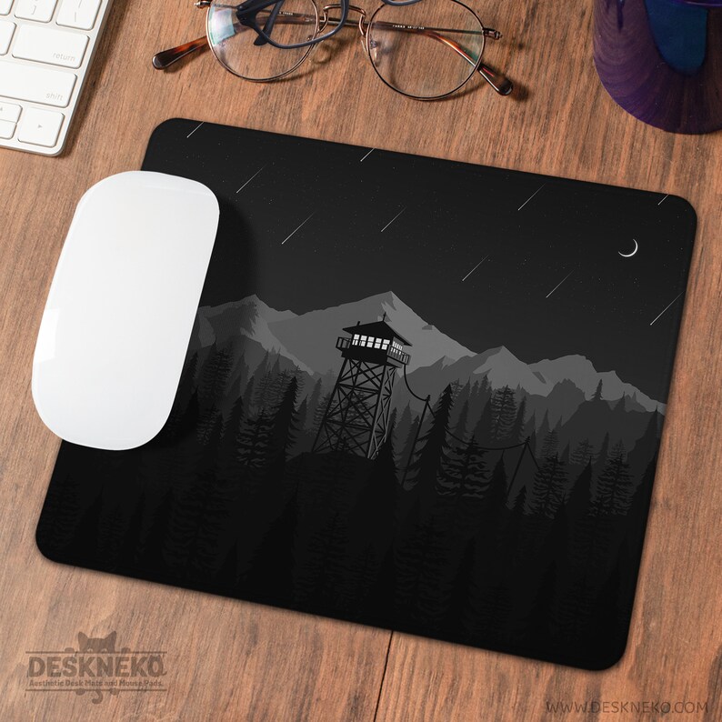 May include: Black mousepad with a mountain and forest design. The illustration features a fire watchtower, trees, mountains, a crescent moon, and shooting stars. A white computer mouse is on the left. The brand name "DESKNEKO" is at the bottom.