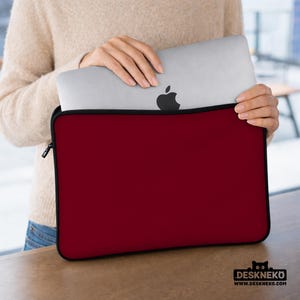 May include: A burgundy laptop sleeve with a black zipper and trim. A silver laptop with the Apple logo is partially visible inside the sleeve. The sleeve is held by a person. The logo for Deskneko is in the bottom right corner.