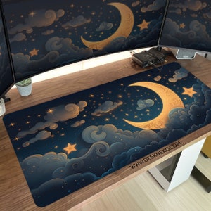 Op de afbeelding: Een rechthoekige bureaumat met een nachthemelontwerp, met een maan, sterren en wolken in blauw, goud en wit. De tekst "WWW.DESKNEKO.COM" staat rechtsonder. De mat ligt op een houten bureau.