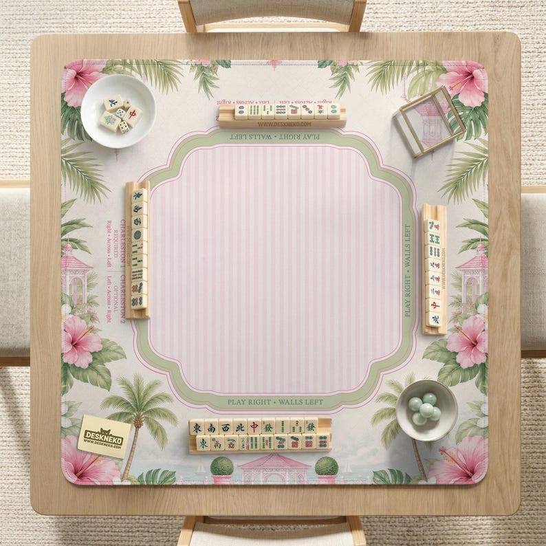 May include: A square mahjong mat featuring a light pink striped center, surrounded by a border of pink hibiscus flowers, green palm leaves, and small gazebos. The mat includes wooden tile racks, a white bowl with tiles, and a small bowl with green dice.