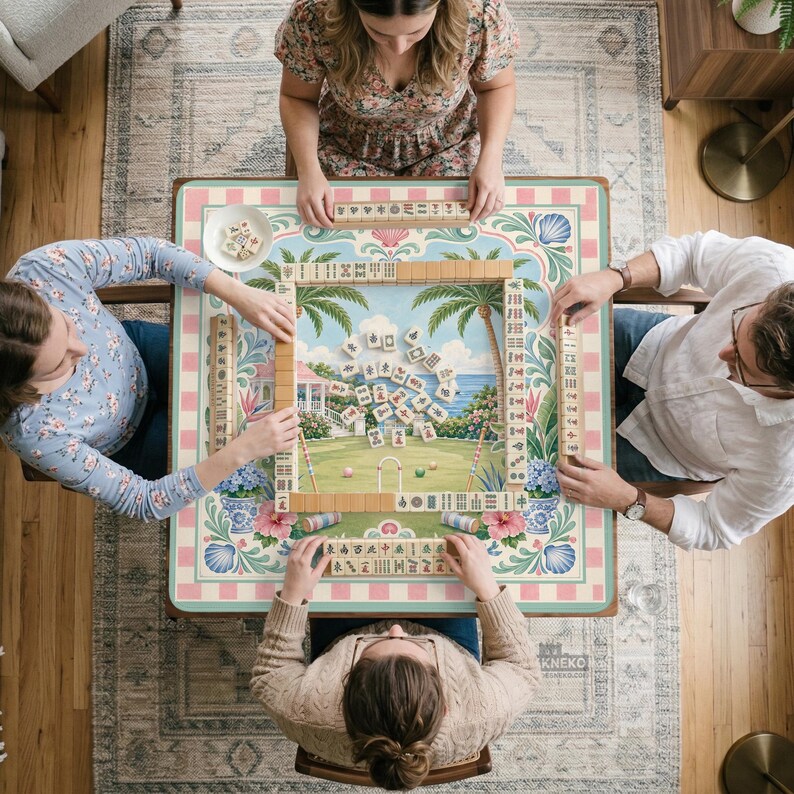 May include: Overhead view of a group playing mahjong. The game board features a tropical scene with a house, palm trees, and ocean. The board is surrounded by pink and blue floral patterns. Players are seated around the table, interacting with the tiles.
