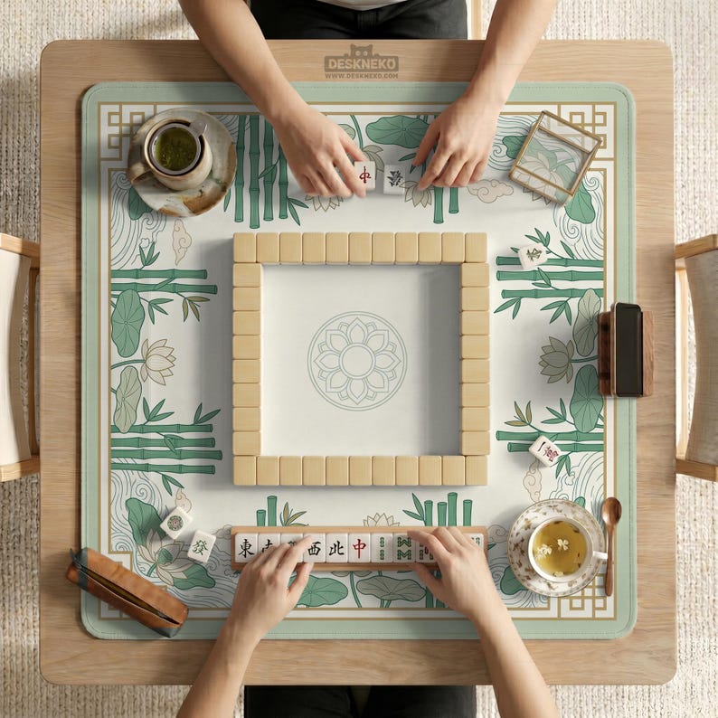 Puede incluir: Vista a&eacute;rea de un juego de mahjong en curso sobre una mesa de madera. El tablero del juego presenta un dise&ntilde;o floral y de bamb&uacute; en verde y blanco. Las fichas de mahjong beige est&aacute;n dispuestas y dos personas est&aacute;n jugando. El texto "DESKNEKO" es visible.