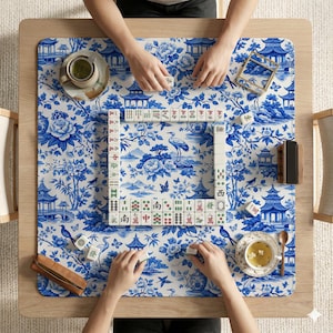 May include: A top-down view of a Mahjong game in progress. The game is set on a wooden table with a blue and white floral patterned mat. Players are arranging tiles, with tea and accessories visible.