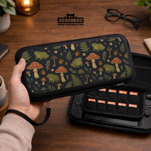 May include: Black case with a whimsical pattern of green frogs, red-capped mushrooms, keys, and acorns. The case is designed for a gaming console and features the text "DESKNEKO" and "WWW.DESKNEKO.COM".