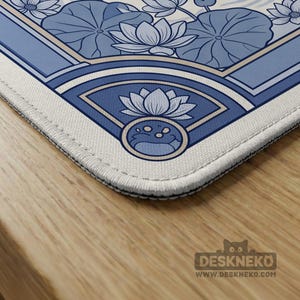 May include: A blue and white desk mat featuring a lotus flower design. The mat has a detailed illustration of lotus flowers and leaves, with a fan-shaped design in the corner. The mat has a white border and the Deskneko logo.
