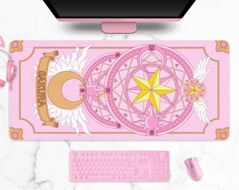 May include: Pink rectangular desk mat with a detailed anime-style design featuring a sun, moon, stars, and text that reads "SAKURA". A pink keyboard, mouse, and headphones are also visible.