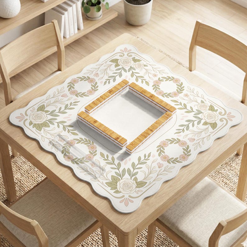May include: A square mahjong mat with a scalloped edge featuring a white background decorated with light green and soft pink floral rose patterns and circular wreaths. In the center sits a square wooden mahjong tile rack set.
