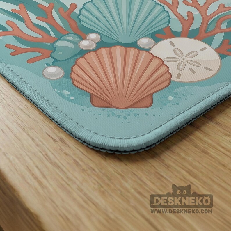 May include: A rectangular desk mat featuring a light blue background with a printed illustration of coral, white pearls, a large teal seashell, a smaller peach-colored seashell, and a sand dollar. The mat has stitched edges and the text DESKNEKO.