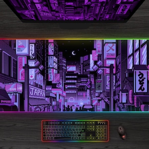 May include: A large rectangular mouse pad featuring a pixelated cityscape in shades of purple and black, with Japanese text and neon signs. The pad has a glowing rainbow edge, and a keyboard and mouse are visible below.