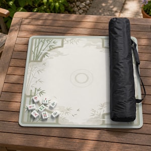 May include: A square mahjong mat featuring a light-colored surface with a green bamboo print and a central circular design. Several white mahjong tiles with red and green characters are scattered on the mat, accompanied by a black carrying case.