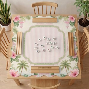 May include: A square mahjong mat with a scalloped edge featuring a pink and white striped center, green borders, and a tropical pattern of palm trees and pink hibiscus flowers. Four wooden tile racks are placed on the mat with mahjong tiles.