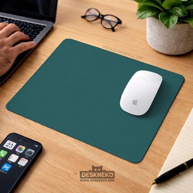 May include: A teal-colored rectangular mousepad with a white Apple mouse. The mousepad is on a wooden desk, with a laptop, glasses, a potted plant, a smartphone, and a pen visible. The mousepad has the text "DESKNEKO" and "WWW.DESKNEKO.COM".