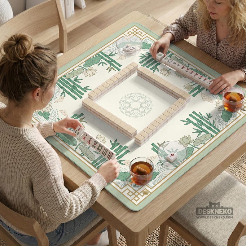 Puede incluir: Una mesa cuadrada de madera con un juego de Mahjong en curso. El tablero del juego presenta una superficie color crema con un dise&ntilde;o floral y de bamb&uacute;. Dos personas est&aacute;n jugando, sosteniendo fichas y tazas de t&eacute;. La mesa tiene el texto "DESKNEKO".