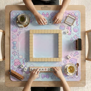 May include: A wooden mahjong set with beige tiles is arranged on a pastel-colored mat with sea creature and bow illustrations. The mat is on a light wood table. The text "DESKNEKO" is visible on the mat.