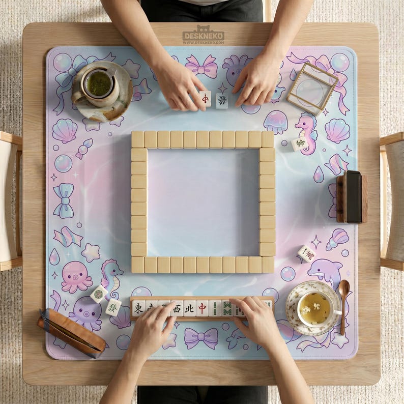 May include: A wooden mahjong set with beige tiles is arranged on a pastel-colored mat with sea creature and bow illustrations. The mat is on a light wood table. The text "DESKNEKO" is visible on the mat.