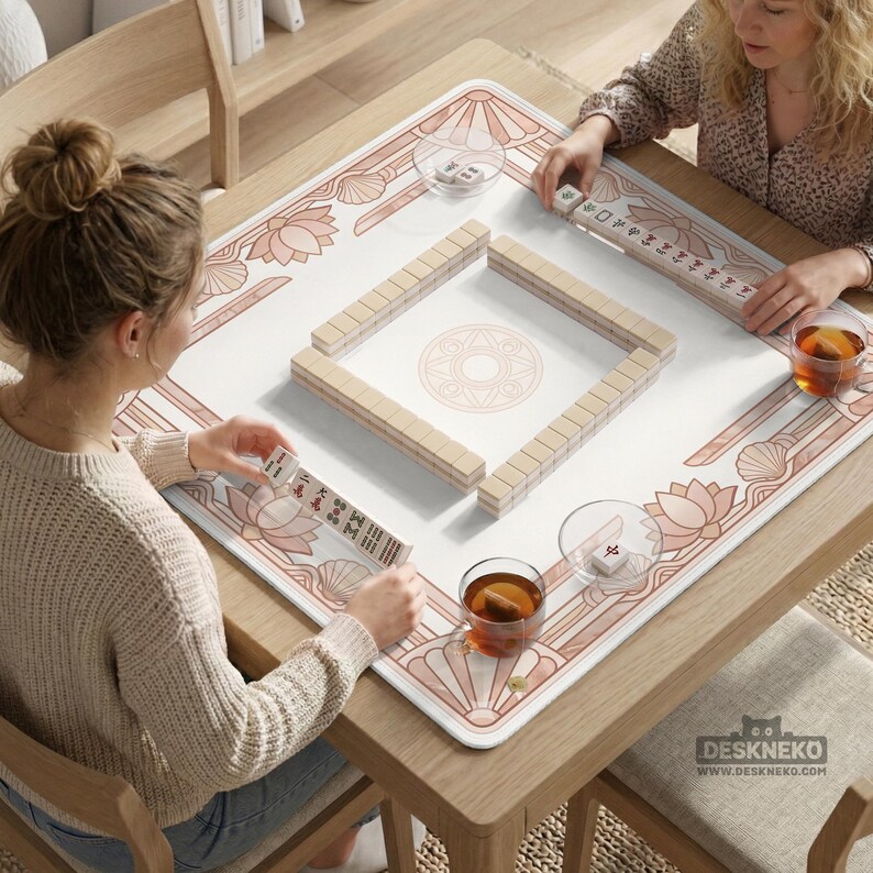 May include: A wooden mahjong table with a white surface and a pink floral design. The table is set for a game, with tiles arranged in a square and two players seated around it. The table has a logo that says DESKNEKO.