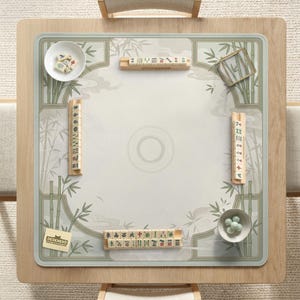 May include: A square mahjong mat featuring a light beige surface with green bamboo illustrations and a central circular motif. The mat includes four wooden tile racks, a white bowl with mahjong tiles, a bowl with green glass beads, and a small card.