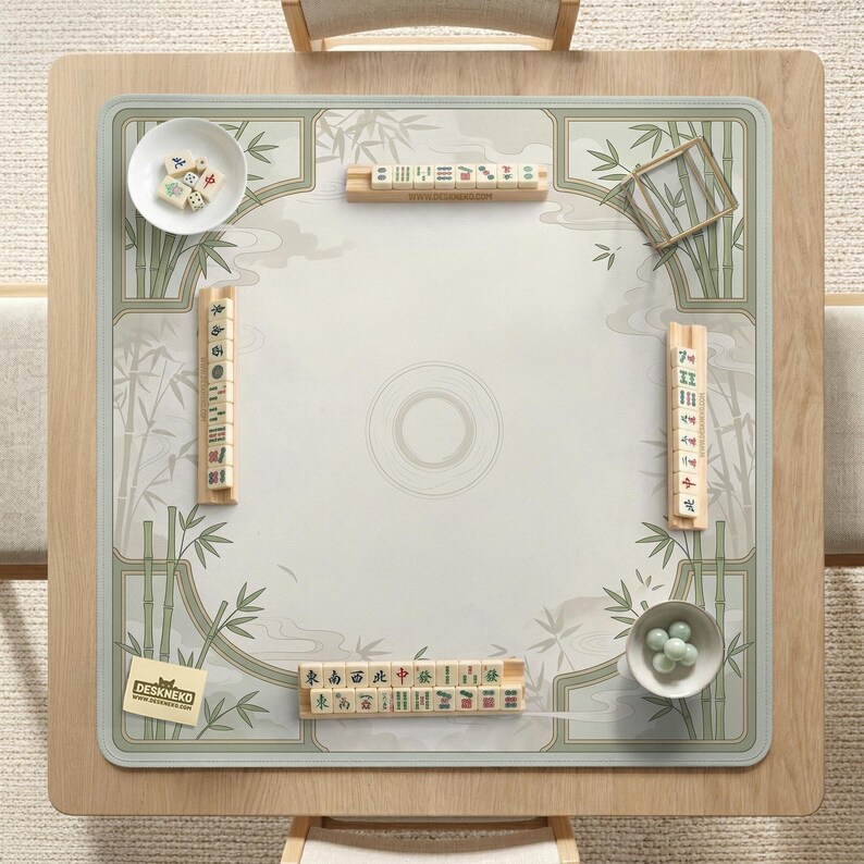 May include: A square mahjong mat featuring a light beige surface with green bamboo illustrations and a central circular motif. The mat includes four wooden tile racks, a white bowl with mahjong tiles, a bowl with green glass beads, and a small card.