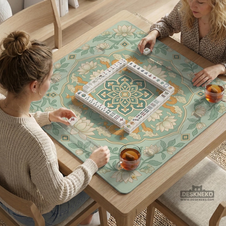 May include: Two people playing Mahjong on a wooden table. The table is covered with a green and beige floral patterned mat. The Mahjong tiles are white with black and red markings. The table also has two cups of tea.