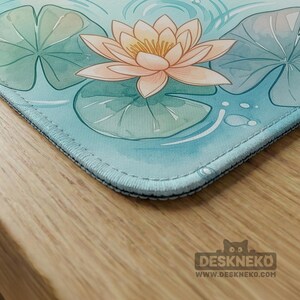 May include: A rectangular desk mat featuring a watercolor illustration of a light peach lotus flower surrounded by green lily pads on a light blue water surface with ripples. The bottom right corner displays the text DESKNEKO WWW.DESKNEKO.COM.
