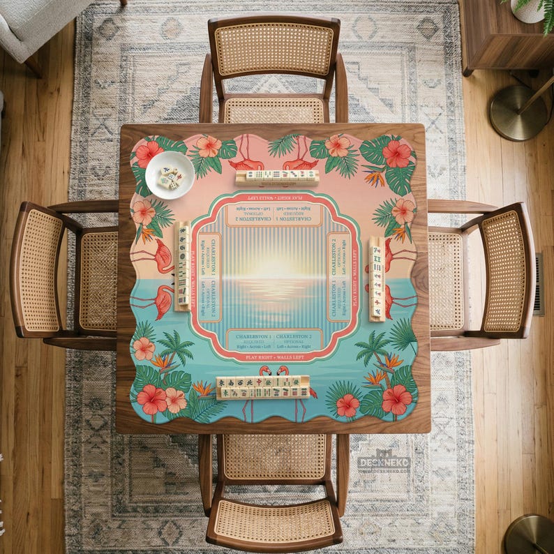 May include: A square mahjong mat featuring a tropical sunset design with pink flamingos, green palm leaves, and red hibiscus flowers. The center displays game instructions and charts, surrounded by four wooden tile racks and a small white bowl.