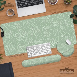 Op de afbeelding: Een bureau-opstelling met een groene muismat, polssteun en muismat, allemaal met een wit monstera-bladpatroon. Een wit toetsenbord, muis en laptop zijn ook aanwezig. De website www.deskneko.com is zichtbaar.